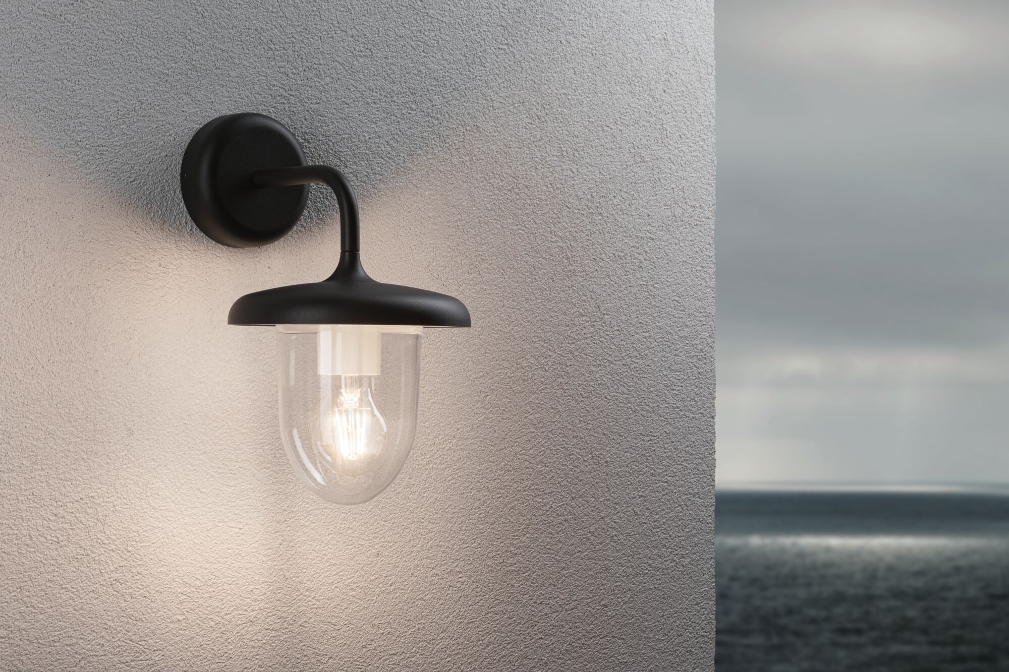 Sort udendørs væglampe med klart glas og synlig pære på hvid væg med havudsigt