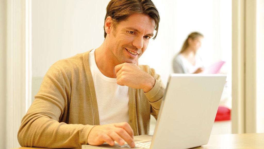 Mann in beigem Cardigan arbeitet konzentriert an weißem Laptop in hellem Raum mit Frau im Hintergrund.