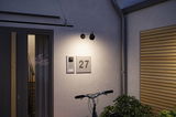 Modern black metal outdoor wall light illuminating house number 27 and door intercom on grey wall.