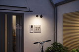 Modern black metal outdoor wall light illuminating house number 27 and door intercom on grey wall.