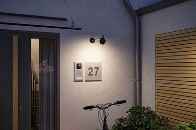 Moderne schwarze Außenwandleuchte aus Metall beleuchtet Hausnummer 27 und Türsprechanlage an grauer Wand.