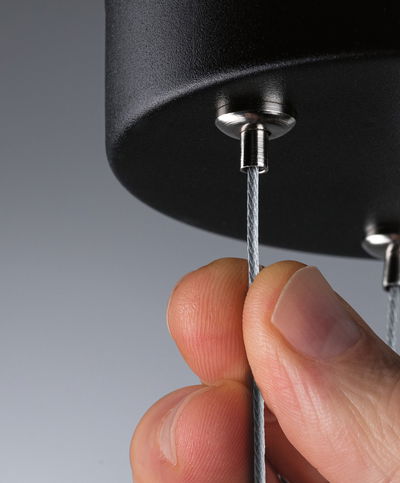 Close-up van een hand die een dun stalen kabel vasthoudt aan een zwarte metalen houder, verlichtingsonderdeel.