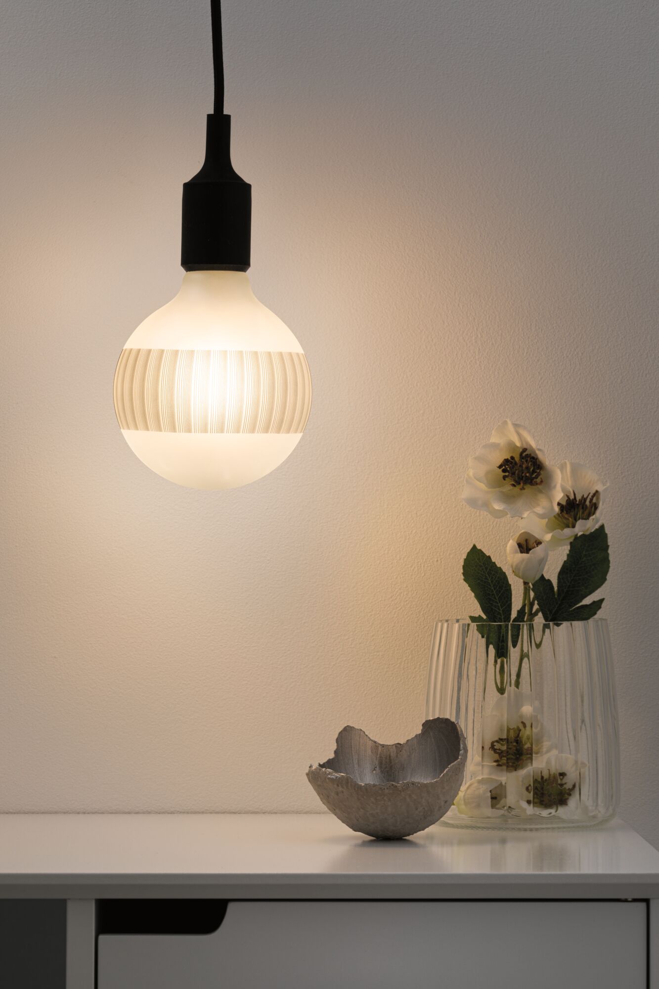 Hangende LED-lamp met zwarte fitting en warm wit licht boven witte tafel met vaas en decoratie.