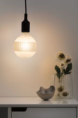 Hanging LED bulb with black socket and warm white light above white table with vase and decoration.