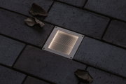 Recessed square stainless steel LED floor light with warm white glow on dark pavement