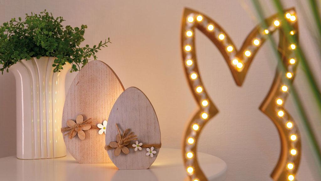 Decorative wooden Easter eggs with flower details beside illuminated bunny frame and white vase with green plant