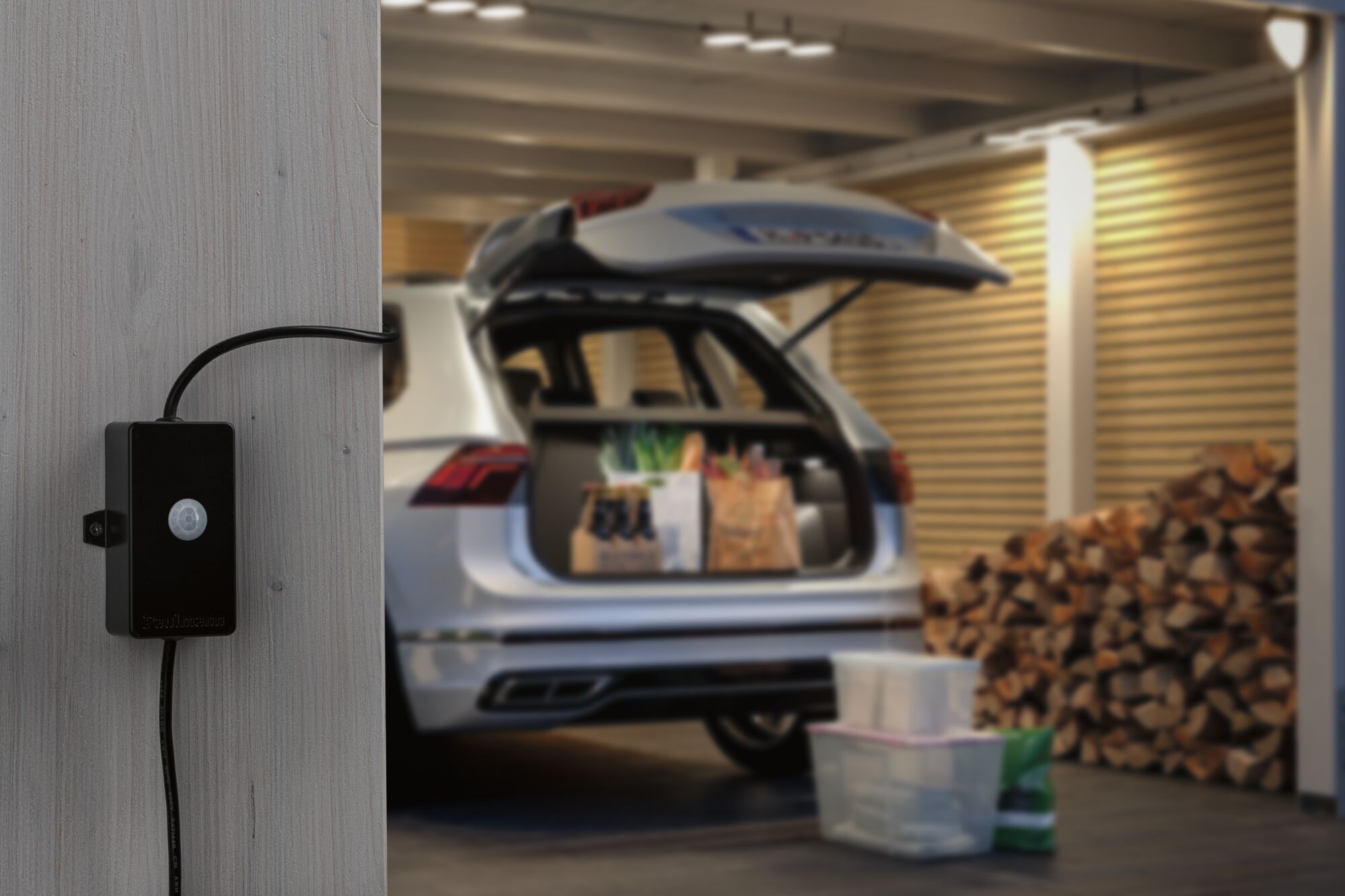 Zwarte kunststof bewegingssensor aan houten wand in garage met open kofferbak en gestapeld brandhout.