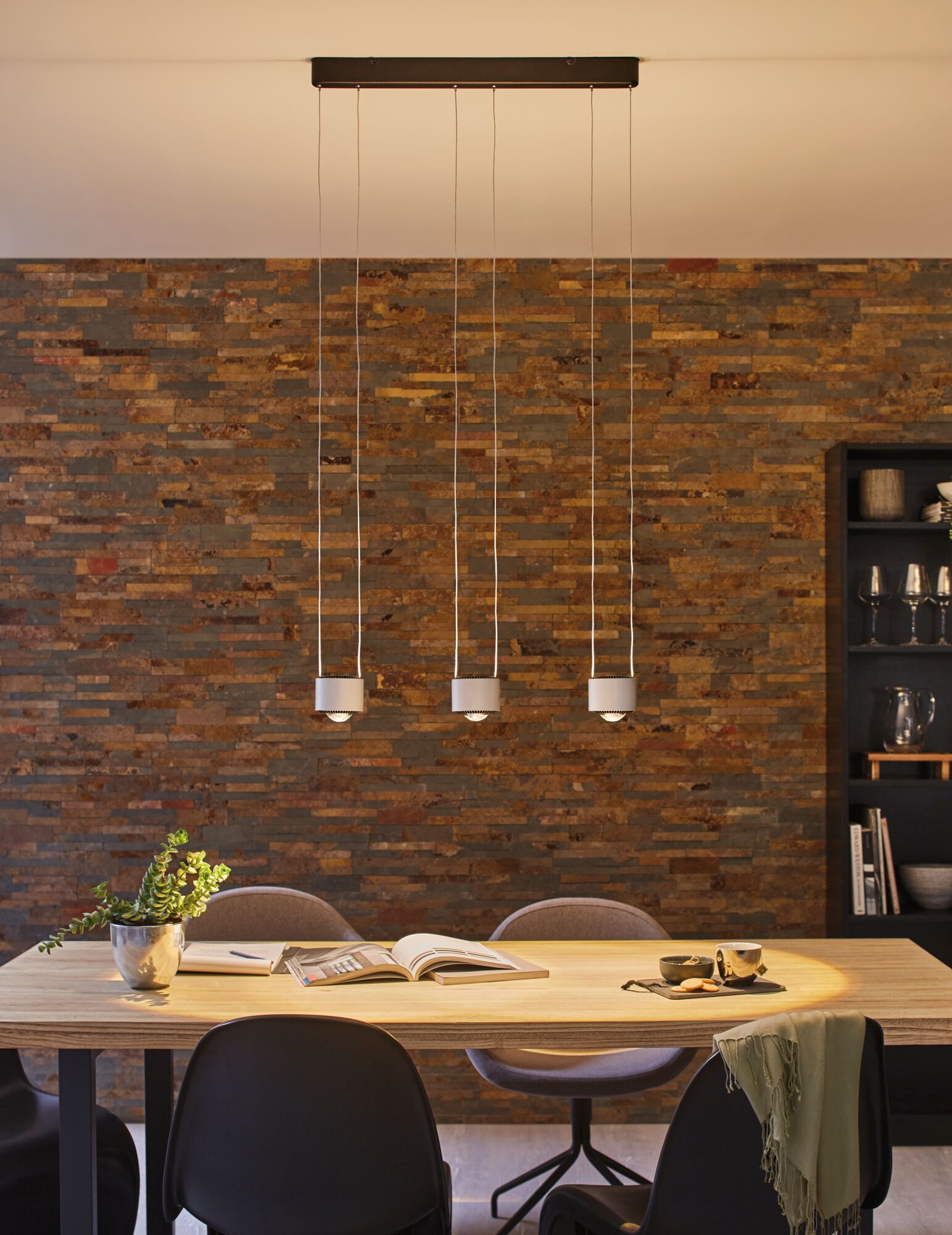 Modern white three-light pendant lamp above wooden table with books and plant in dining room