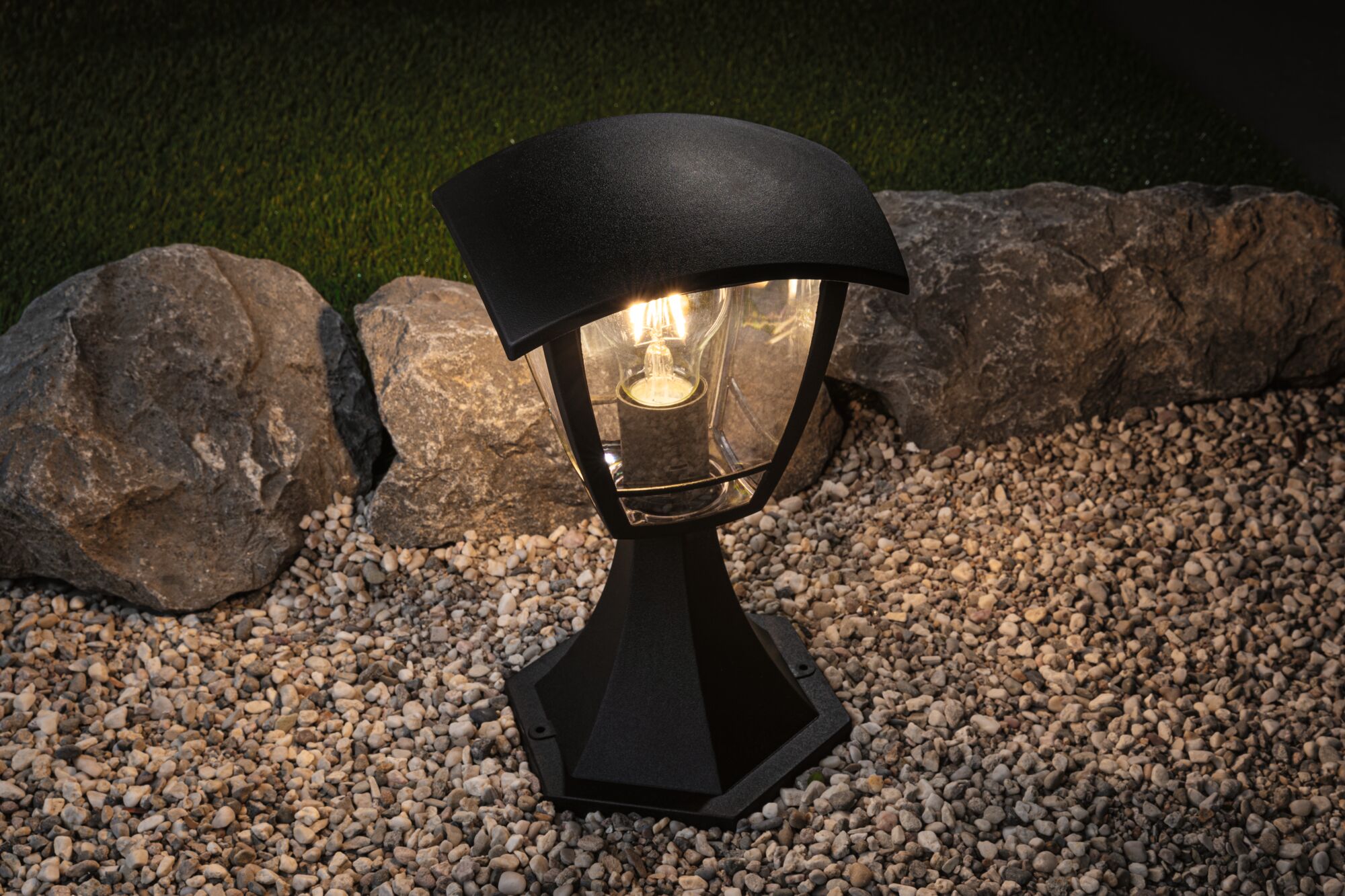 Black outdoor metal lantern with glass and warm white light on gravel and stones in garden