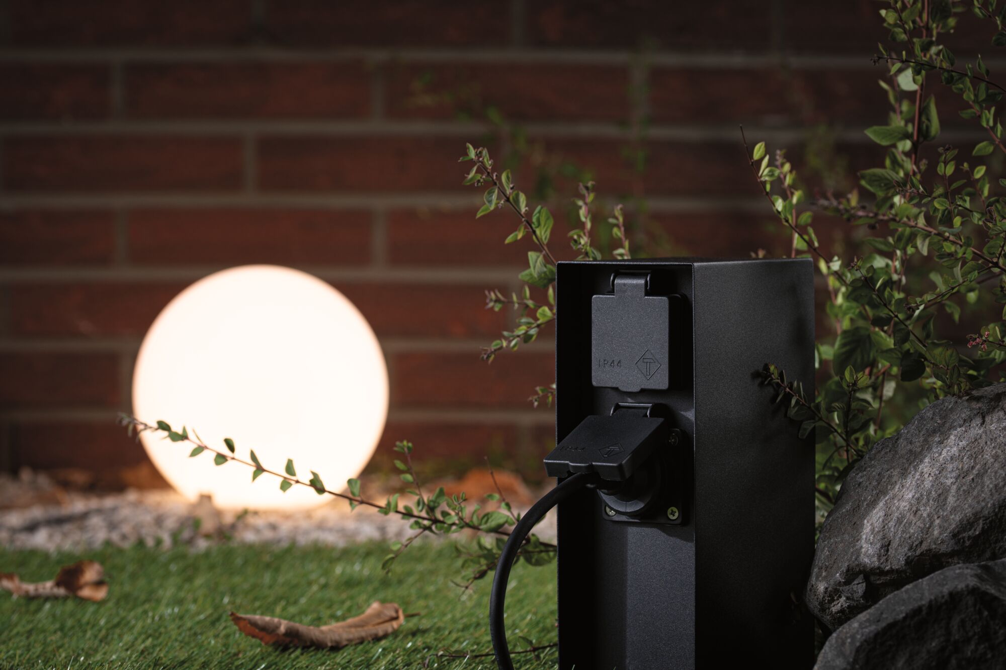 Prise extérieure noire IP44 devant une sphère lumineuse dans un jardin avec pierre et plantes.