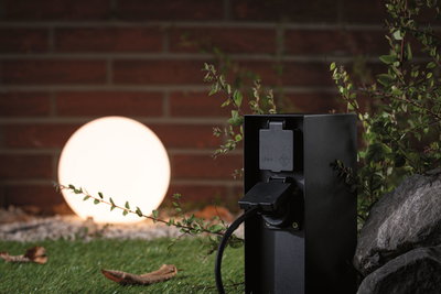 Prise extérieure noire IP44 devant une sphère lumineuse dans un jardin avec pierre et plantes.