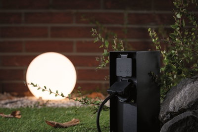 Black outdoor socket with IP44 protection near glowing garden sphere with stone and plants.