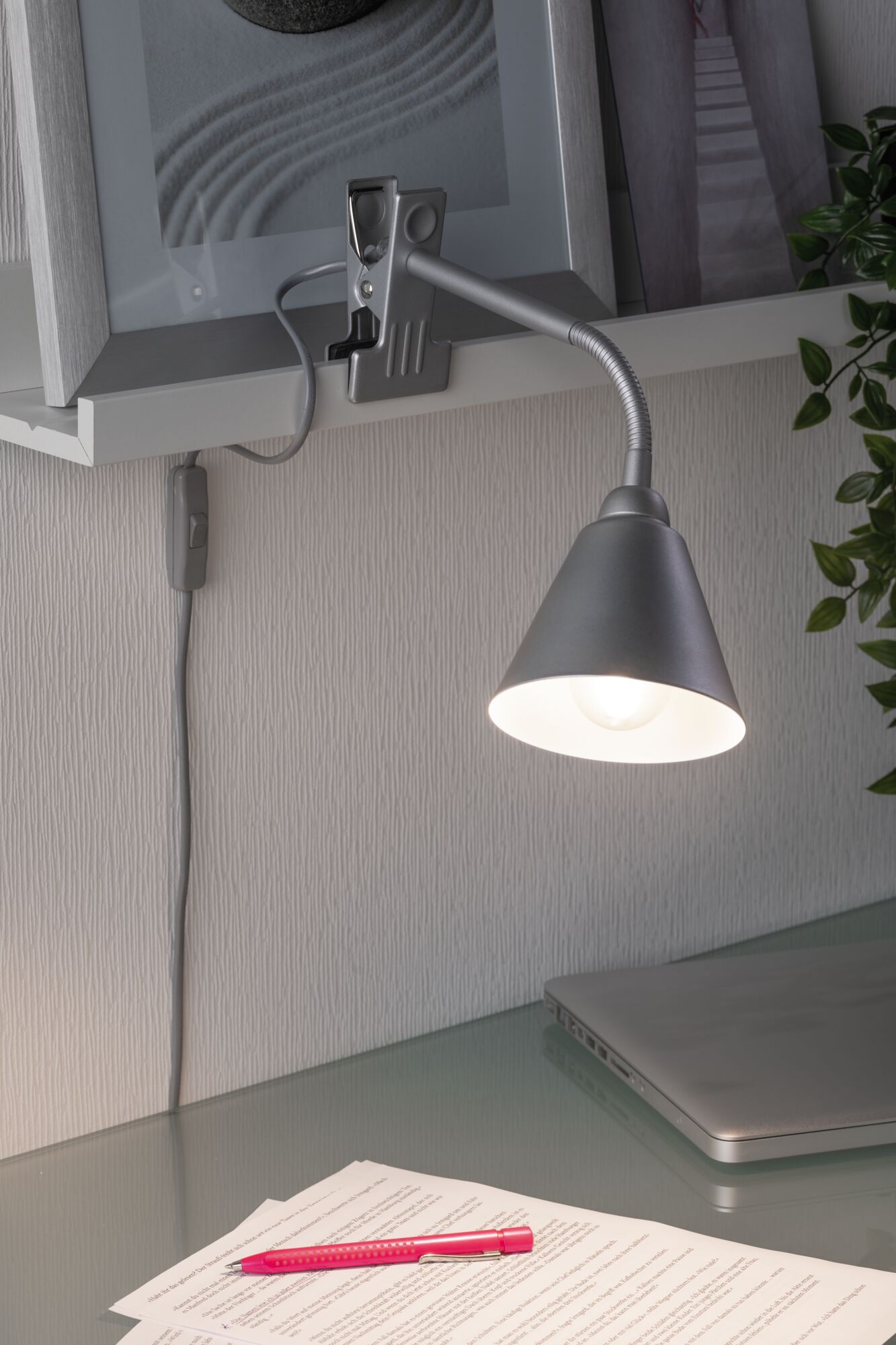 Graue Klemmlampe mit flexiblem Arm an Regal, beleuchtet Schreibtisch mit Laptop und Dokumenten.