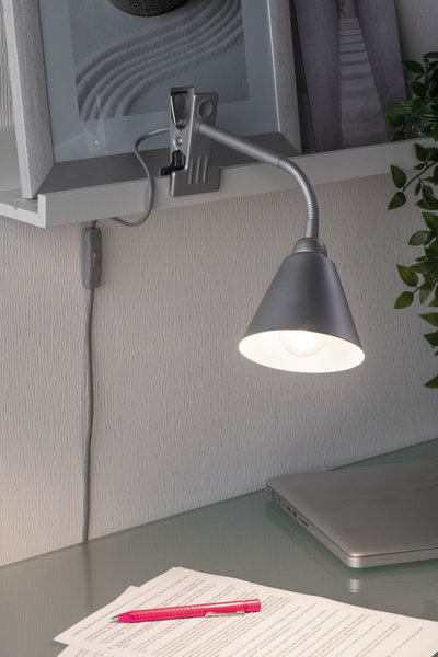 Graue Klemmlampe mit flexiblem Arm an Regal, beleuchtet Schreibtisch mit Laptop und Dokumenten.