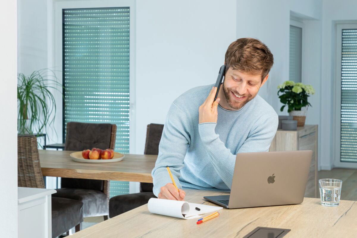 Man in lichtblauwe trui belt en maakt aantekeningen bij laptop in moderne eetkamer met houten tafel