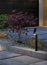 Black modern metal LED pathway light illuminating garden path with gravel and blooming magnolia shrub