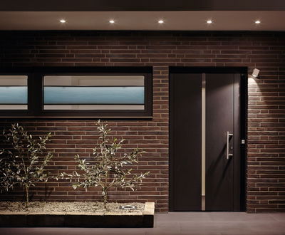 Modern house facade with brown bricks, black door, and energy-saving LED ceiling spotlights outside.