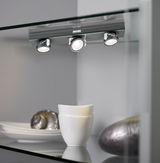 Drie verstelbare matte grijze LED-spotlights op glazen plank met wit servies en decoratie.