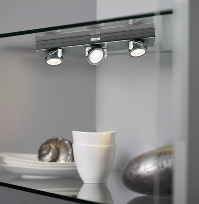 Three adjustable matte grey LED spotlights on glass shelf with white dishes and decorative items.