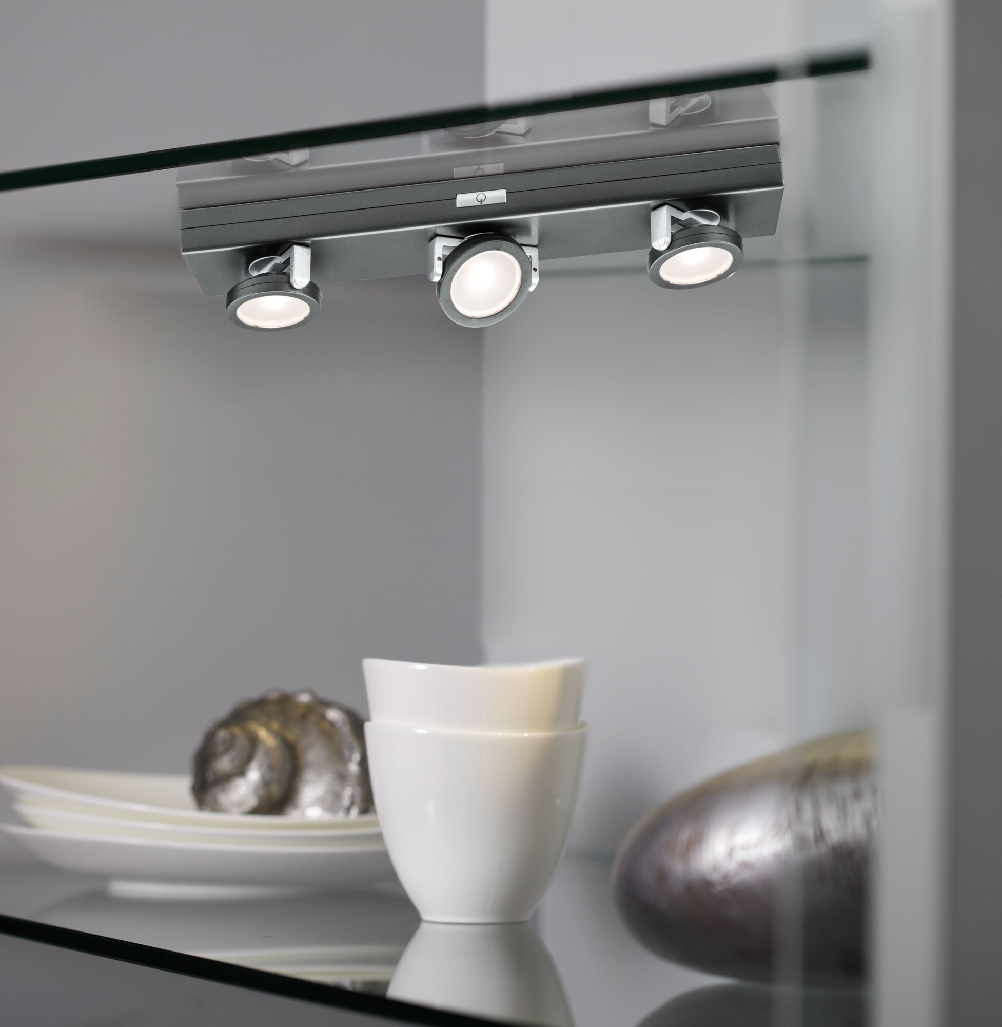 Three adjustable matte grey LED spotlights on glass shelf with white dishes and decorative items.