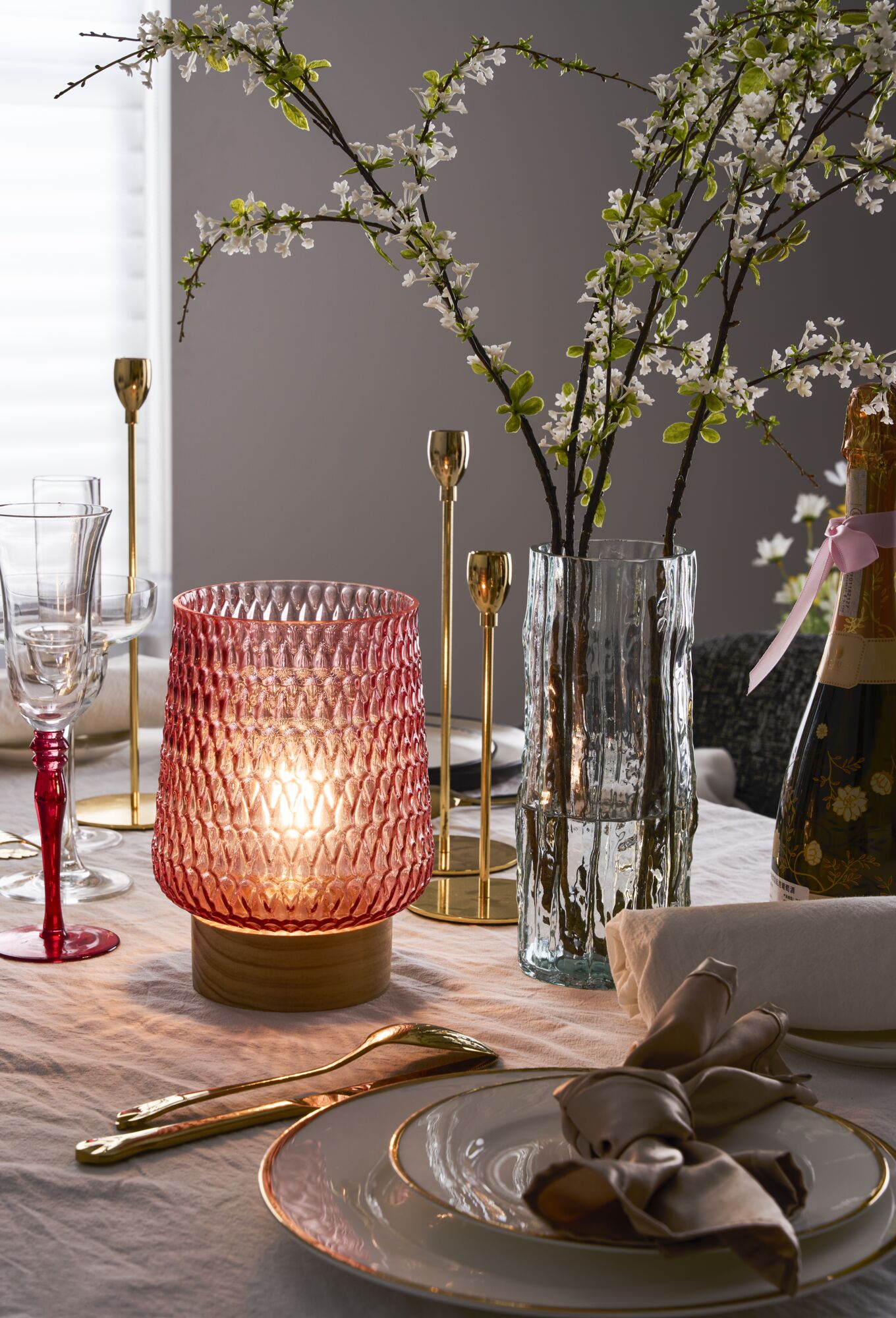 Pink glass lamp with wooden base and clear vase with white blossoms on table set with gold cutlery