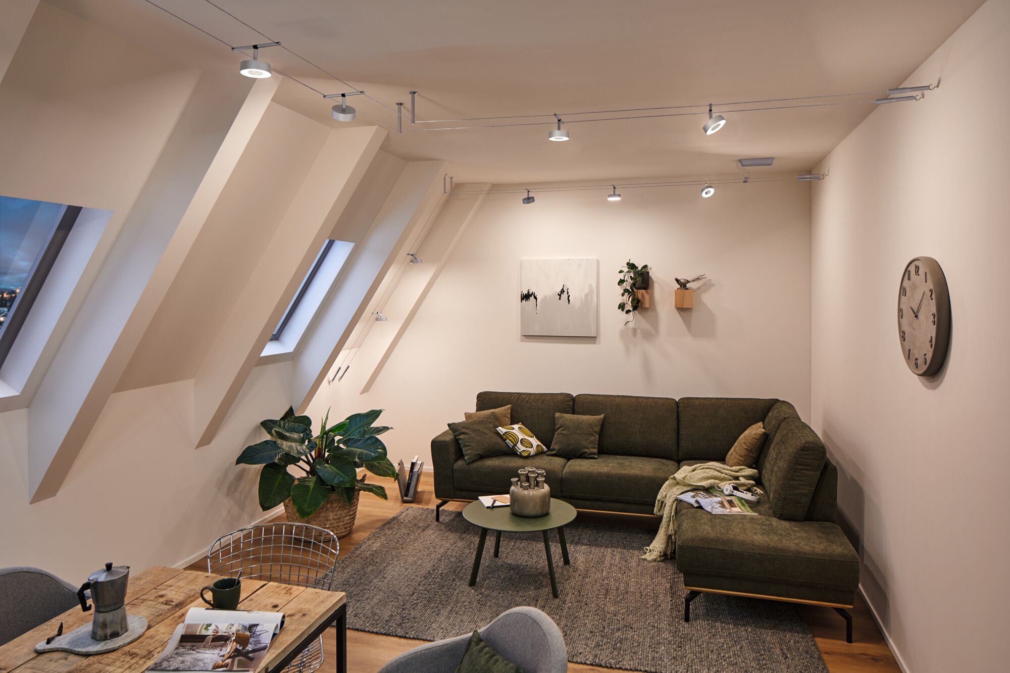 Modernes Wohnzimmer mit grünem Ecksofa, grauem Teppich und Paulmann LED-Seilstrahlern an der Decke.