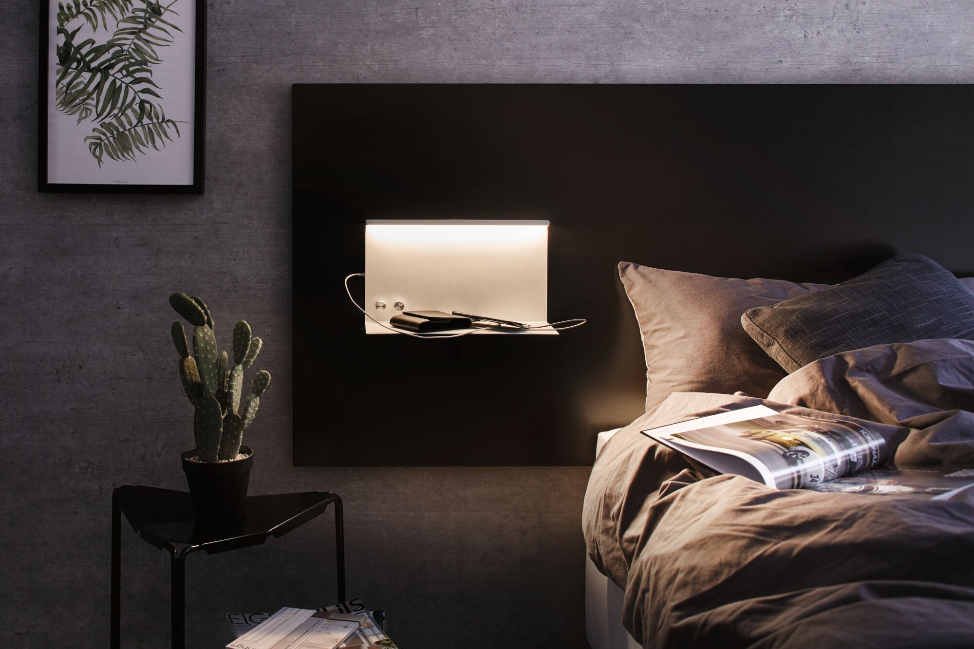 Modern white LED wall light with shelf on black headboard in bedroom with grey wall and cozy bedding.