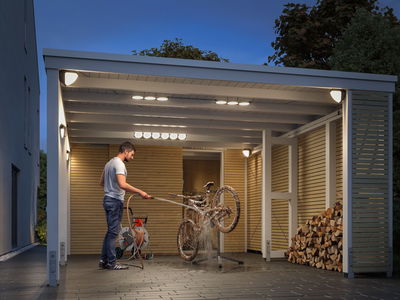 Lit carport with white LED ceiling and wall lights, man cleaning muddy bike with hose in evening.