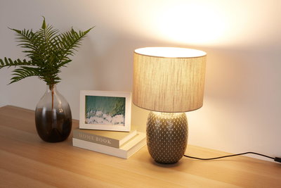 Bordlampe med mønstret keramisk fod og beige stofskærm på træbord med dekoration.