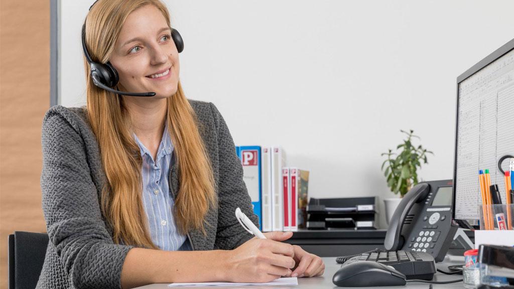 Junge Frau mit Headset am Schreibtisch schreibt Notizen, Büroarbeitsplatz mit Telefon und Computer.