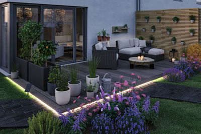 Moderne Terrasse mit LED-Bodenbeleuchtung, grauem Polyrattan-Sitzmöbel und bunten Blumen im Garten.