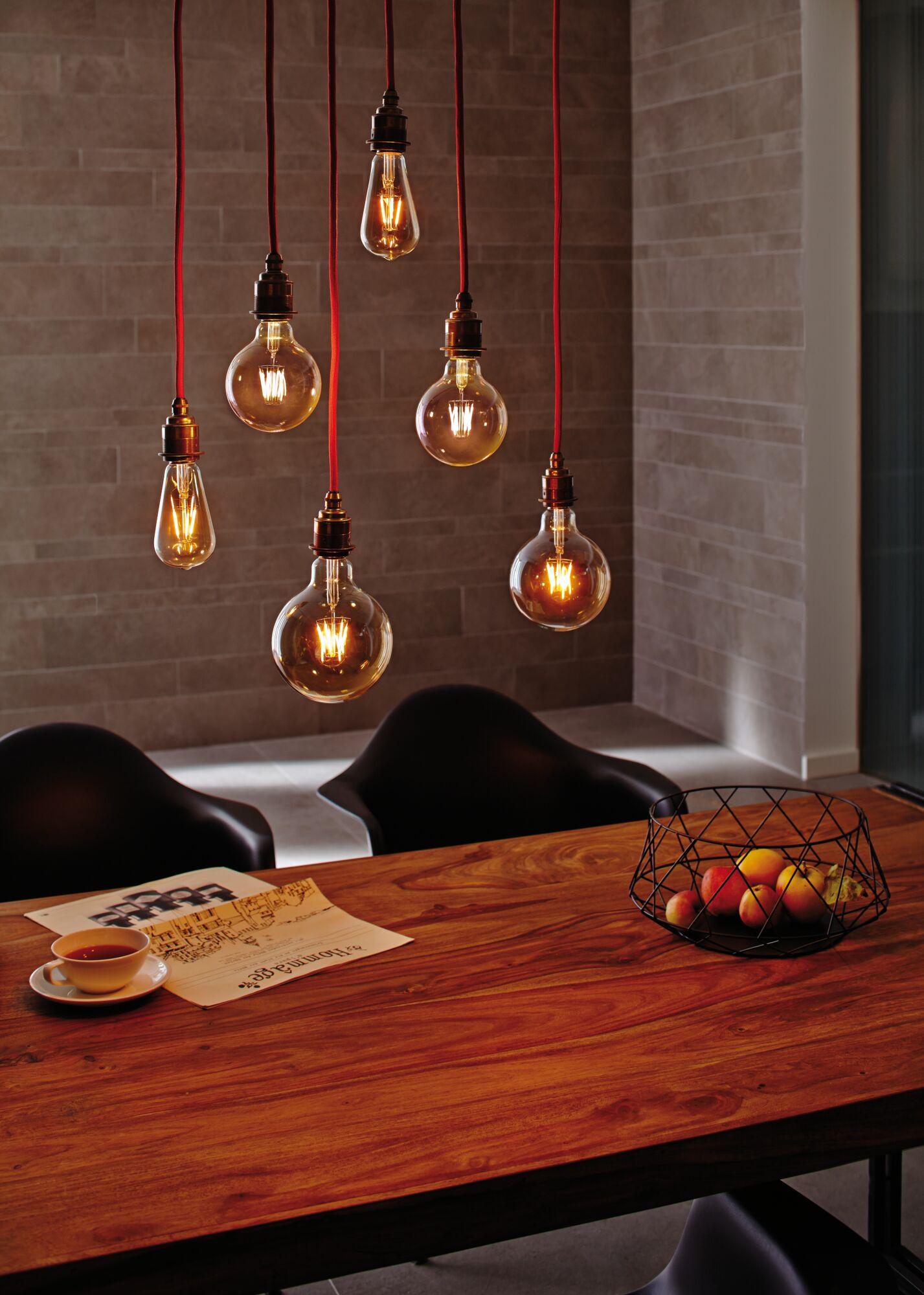 Six hanging light bulbs with red cords above wooden table with fruit basket and newspaper in dining room
