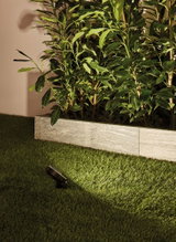 Zwarte LED-tuinschijnwerper op groen gras die planten bij een grijze stenen muur verlicht