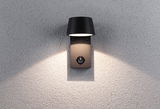 Zwarte metalen buitenwandlamp met bewegingssensor en warm wit licht op grijze muur