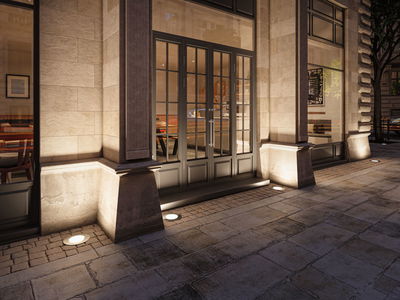 Außenbeleuchtung mit runden Bodeneinbauleuchten in warmweiß vor Sandsteinfassade und Glastür.