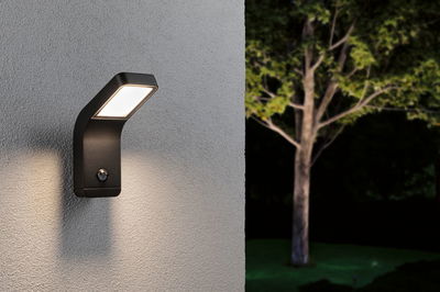 Black LED outdoor wall light with motion sensor mounted on grey wall illuminating garden at night