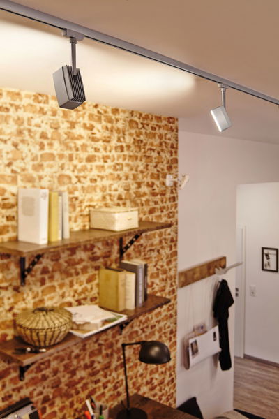 Moderner silberner LED-Schienenstrahler an Decke über Regalwand mit Backsteinmuster und Arbeitsbereich