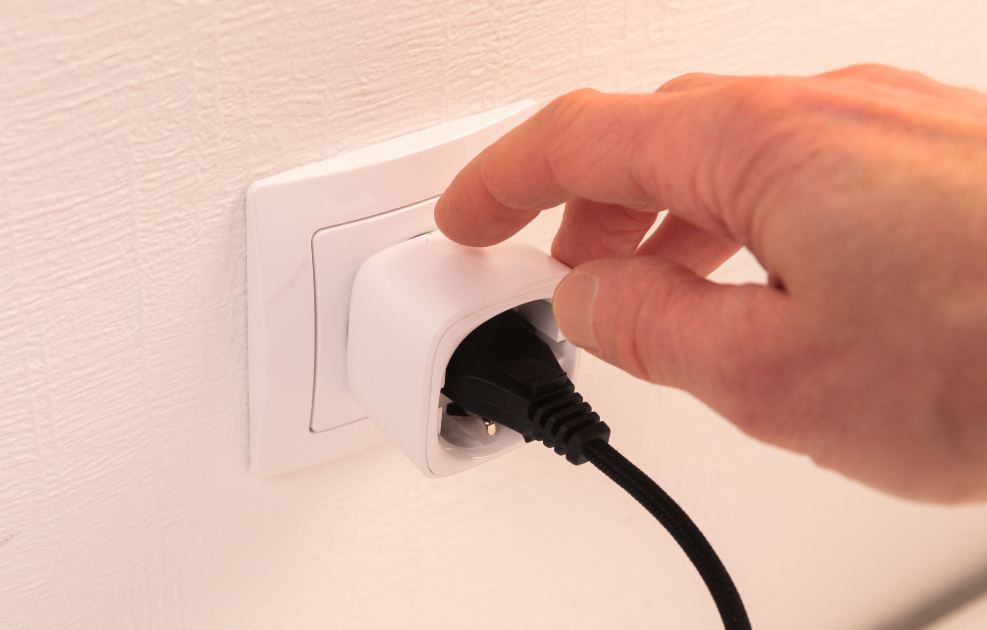 Hand operating white power socket adapter with black cable on textured wall for electrical connection