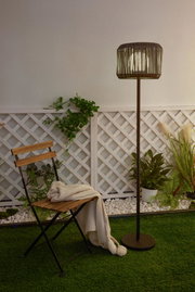 Outdoor floor lamp with brown metal frame and woven shade beside wooden chair on green grass