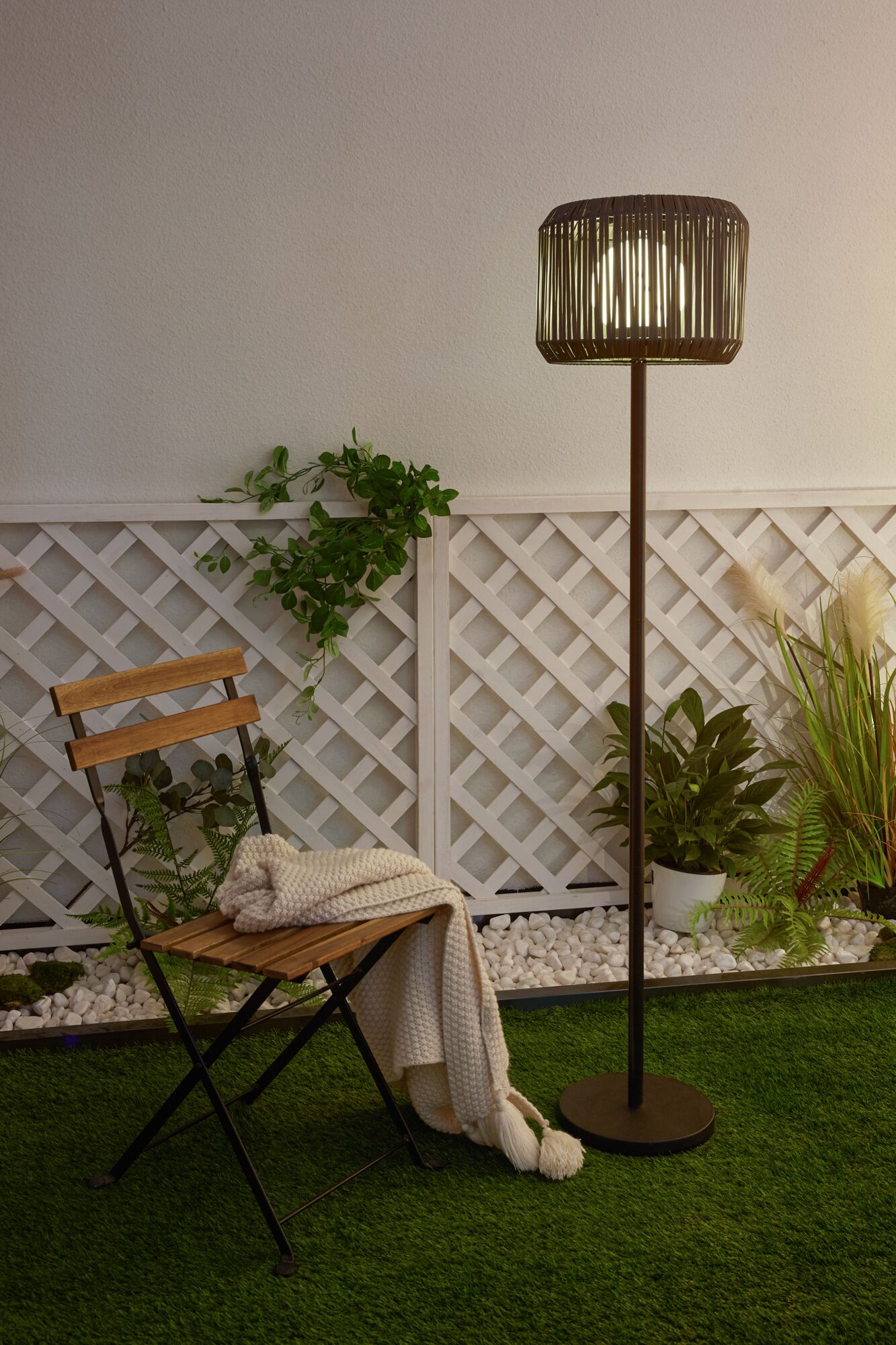 Outdoor floor lamp with brown metal frame and woven shade beside wooden chair on green grass
