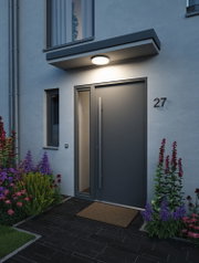 Round white LED ceiling light in plastic above modern house entrance with dark door and garden plants
