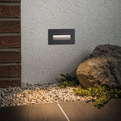 Rechthoekige zwarte metalen LED-wandlamp op witte muur met warm buitenlicht en tuinstenen