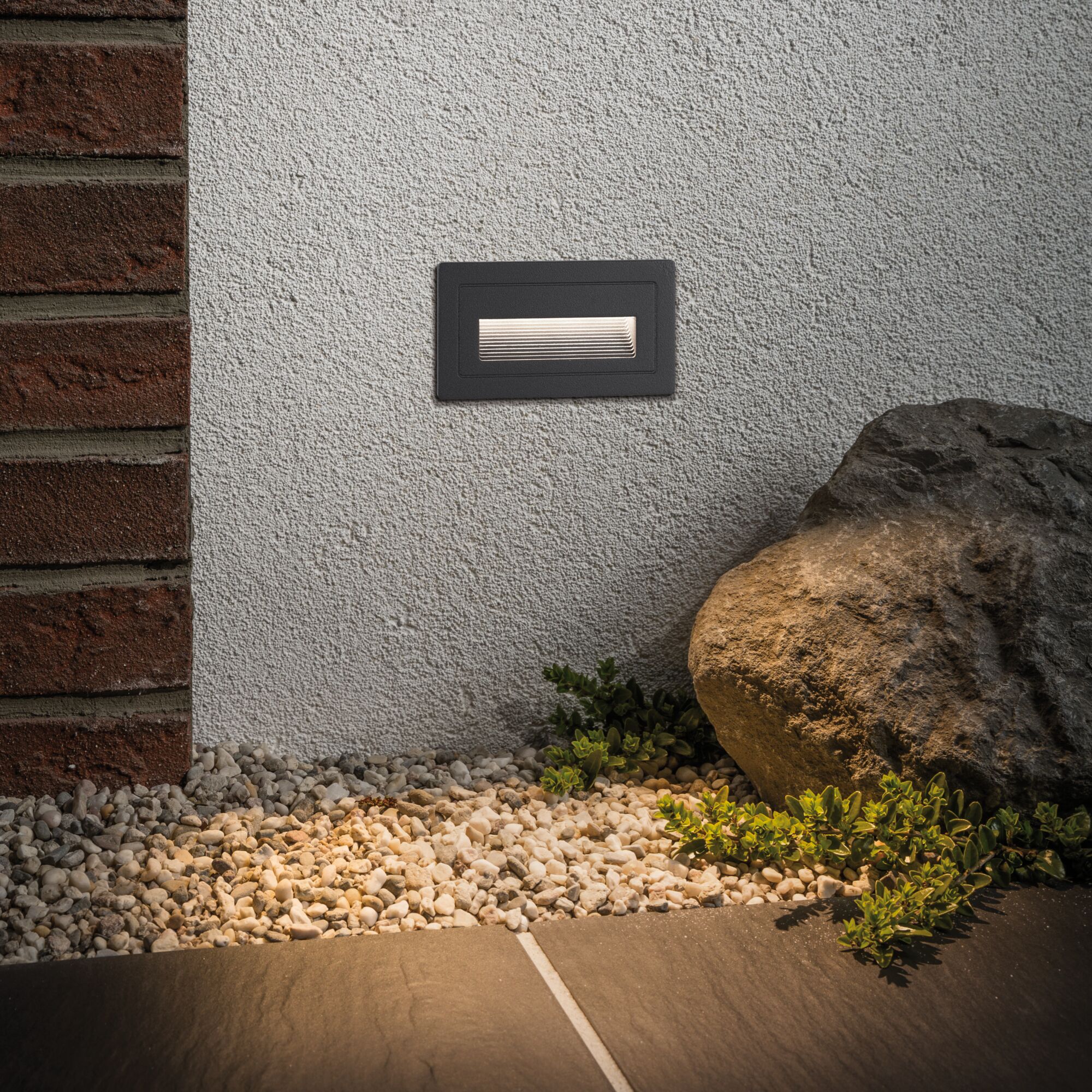 Rectangular black metal LED wall light on white wall with warm outdoor lighting and garden stones