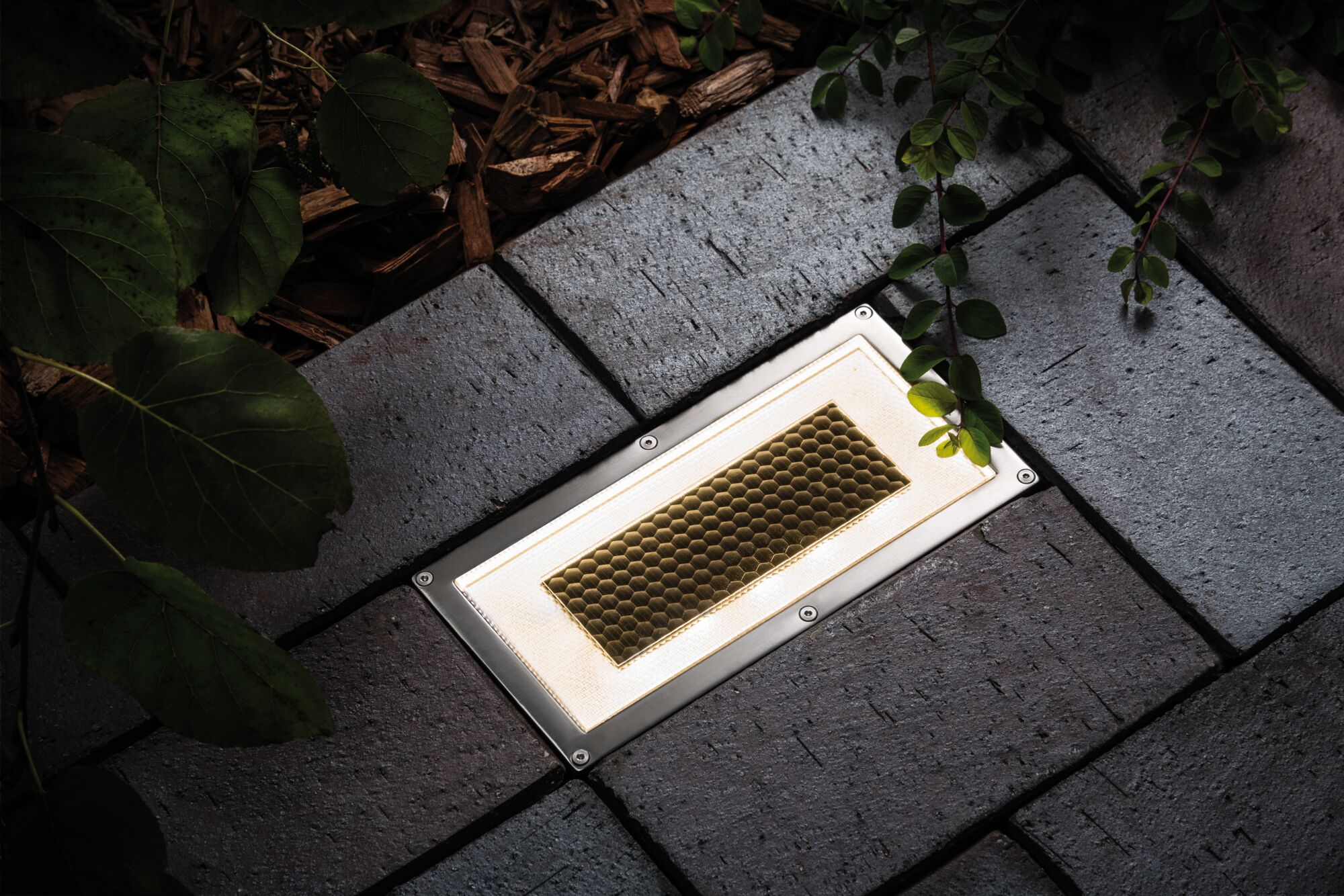 Eingelassene LED-Bodenleuchte mit Edelstahlrahmen und honigwabenförmigem Glas in grauem Pflaster.