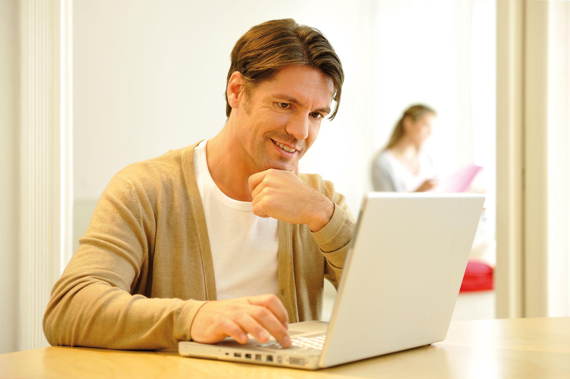 Homme en cardigan beige travaillant concentré sur un ordinateur portable argenté à une table en bois clair