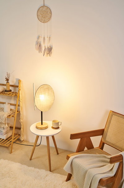 Modern table lamp with wooden base and beige fabric shade on white side table next to wooden chair with blanket