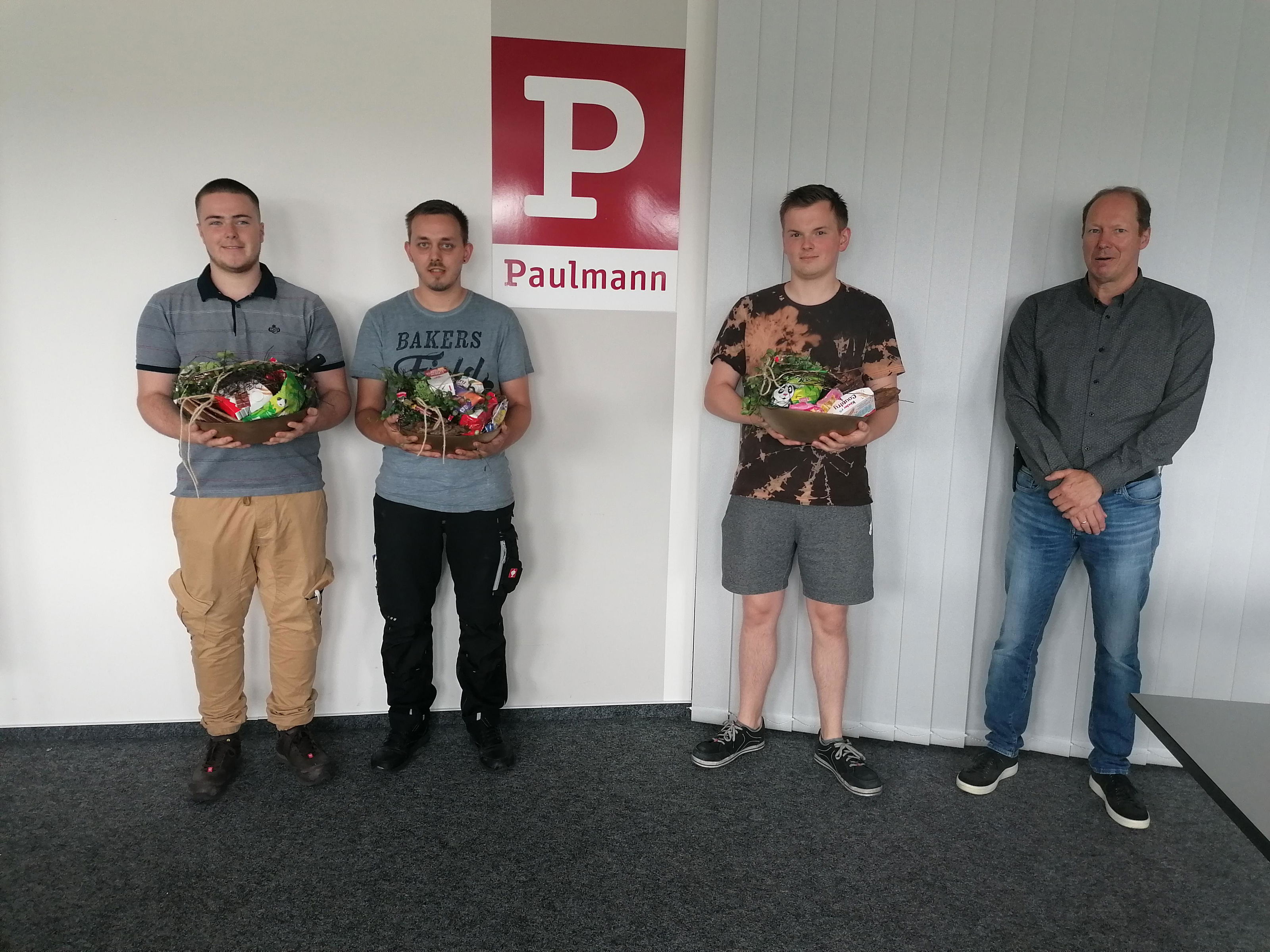 Three young men holding gift baskets in front of Paulmann logo in office, representing training and career.