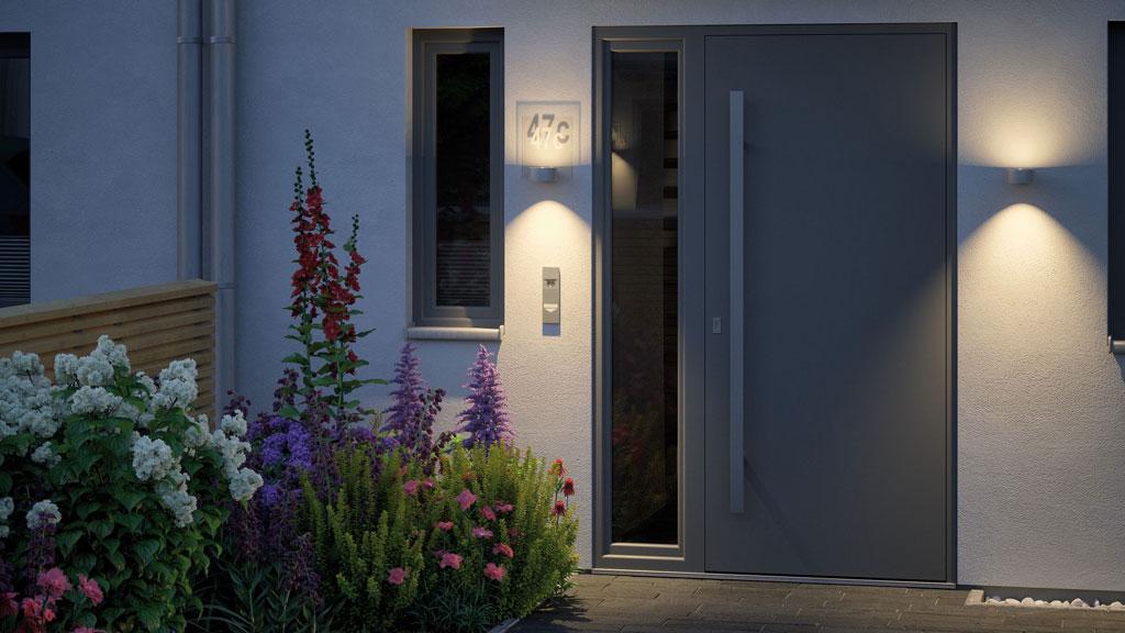 Moderne buitenverlichting met warm wit licht bij grijze voordeur en kleurrijk bloemenperk.