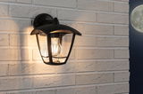 Zwarte metalen buitenwandlamp met helder glas en warm wit LED-licht op lichte bakstenen muur