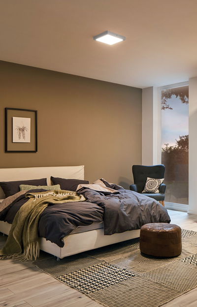 Chambre moderne avec plafonnier carré blanc, lit confortable avec linge de lit foncé et pouf en cuir marron